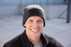 A black beanie with a patch, worn by a smiling man against a modern outdoor backdrop.