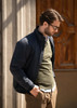 A man wearing a navy jacket over an olive sweater, standing outside next to a wooden door. The jacket has a logo.