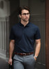 A navy polo shirt worn by a man with glasses, paired with grey trousers and a belt, standing in an outdoor setting.