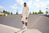 An oversized hooded jacket in beige is worn with matching shorts, set in a sunny skatepark. The model stands on a ramp.