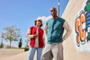 A red and teal varsity jacket worn by two individuals outdoors, with a graffiti background and blue sky above.