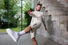 A man in light beige sports shorts and a matching hoodie is performing a kicking motion in a modern interior.