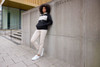 A woman in a colour-block black and cream hoodie leans against a grey wall, wearing beige trousers and white shoes.