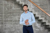 A light blue men's shirt is displayed on a model standing next to a concrete wall and staircase. The shirt has a logo.
