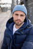 A man wearing a blue fisherman beanie and a navy puffer jacket stands outdoors with a bridge in the background.