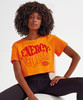 A model wearing an orange crop top with red text, featuring a logo, paired with black leggings.