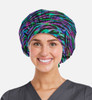 A printed unisex bouffant cap in a vibrant purple and green pattern, worn by a smiling individual in scrubs.