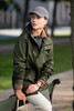 A green unisex jacket is worn by a woman sitting on a bench, paired with a grey cap and neutral trousers.