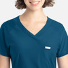 A close-up of a woman wearing a teal Embrace Luna Tuckable Y-Neck Top with a small pocket and branding.
