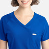 A close-up of a woman's blue Y-neck top with a pocket and branding label. The fabric appears smooth and comfortable.