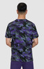 A camouflage print t-shirt in shades of purple, green, and black, viewed from the back. The model is wearing purple pants.