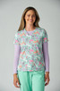 A woman models a colourful scrub top with a floral pattern, featuring shades of pink and green, layered over a long-sleeve top.