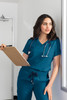 A woman in teal scrubs holds a clipboard and leans against a wall, with a stethoscope around her neck.