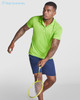 A man wearing a bright green polo shirt and navy shorts holds a badminton racket. The shirt features a logo.