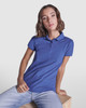 A blue women's premium polo shirt with a collar, featuring a logo, displayed by a model seated on a wooden block.