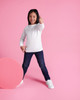 A child poses in a white long-sleeve t-shirt and blue jeans against a pink background, giving a thumbs-up.