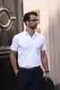 A man in a white polo shirt stands casually with his hands in his pockets, wearing dark trousers and glasses.