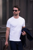 A white men’s T-shirt is displayed, worn by a man carrying a bag and dressed in dark jeans and sunglasses.