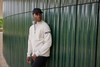 A men's zip jacket in cream, featuring a logo, worn by a model against a textured green wall.