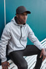 A grey men's sports softshell jacket with a hood, worn by a model seated on a bench, paired with a black cap.
