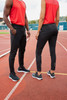 Black track pants worn by two models, paired with red tops and athletic shoes, on an outdoor running track.