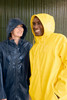 A yellow unisex raincoat and a dark blue raincoat worn by two smiling individuals in the rain. Both jackets have hoods.