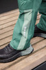 Workwear pants in green with a lighter green panel, featuring reflective details, paired with black footwear on a wooden surface.
