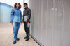 Two models wearing workwear softshell jackets in shades of blue and black, standing against a silver mesh wall.