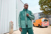 A model wearing a green softshell vest stands outdoors, paired with matching green workwear. A vehicle is visible in the background.