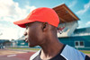 An orange 7 Panel Sports-Cap worn by a person, displayed against a sports facility backdrop.