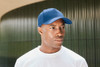 A blue 5 panel laser-cut cap worn by a person, featuring a logo on the front, against a blurred background.