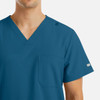 A men's v-neck top in teal with four pockets and a shoulder snap detail. Features branding on the sleeve.