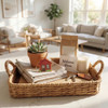 A woven basket holds a plant, folded towels, a candle, a welcome note, keys, and a decorative house piece, all in a sunlit room.