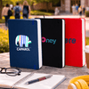 A set of three A5 notebooks in navy, black, and red, placed on a table with a pen and a coffee cup. Each has a logo.