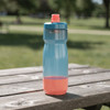 A drink bottle in blue and coral, featuring a screw-top lid and a curved design, resting on a wooden table outdoors.