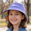 A purple reversible bucket hat with a tie-dye pattern and a logo on the front, worn by a smiling child outdoors.