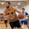 A man wearing a vibrant orange and yellow tank top dribbles a basketball on a gym court, with players in the background.