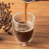 A set of two medium double wall glasses with a hot beverage being poured, surrounded by coffee beans on a wooden surface.