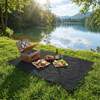 A black camping blanket spread on grass, with a picnic basket, fruit, and sandwiches near a tranquil lake.