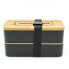A two-layer lunch box in black and natural wood, featuring a secure strap and a branding area on the lid.