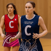 A navy blue netball bib with a white 'C' and logo, worn by a player, alongside a teammate in a red bib.