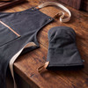 A dark grey canvas apron with light straps and a matching oven mitt, set against a wooden table.