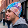 A full colour beanie featuring a vibrant abstract design, worn by a young man in a casual outfit on a city street.