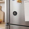 A round black magnetic badge with a logo attached to a stainless steel fridge, alongside a notepad and groceries.