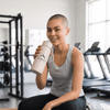 A drink bottle in light grey with a logo, held by a smiling person in a gym setting.