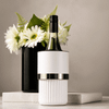 A white wine bottle cooler with a silver band, placed on a marble surface beside a black vase with white flowers.