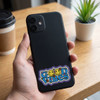 A hand holds a black phone with a colourful "Good Vibes" sticker on its back, placed on a wooden table.