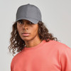 A grey cap worn by a model with curly hair, paired with a pink sweatshirt. The cap features a logo.