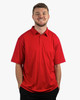 A man wearing a bright red Micro Pique Polo shirt stands smiling, hands in pockets. The shirt has a logo.