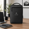 A black soft PU backpack with a logo, placed on a desk alongside a laptop, notebook, and cup.
